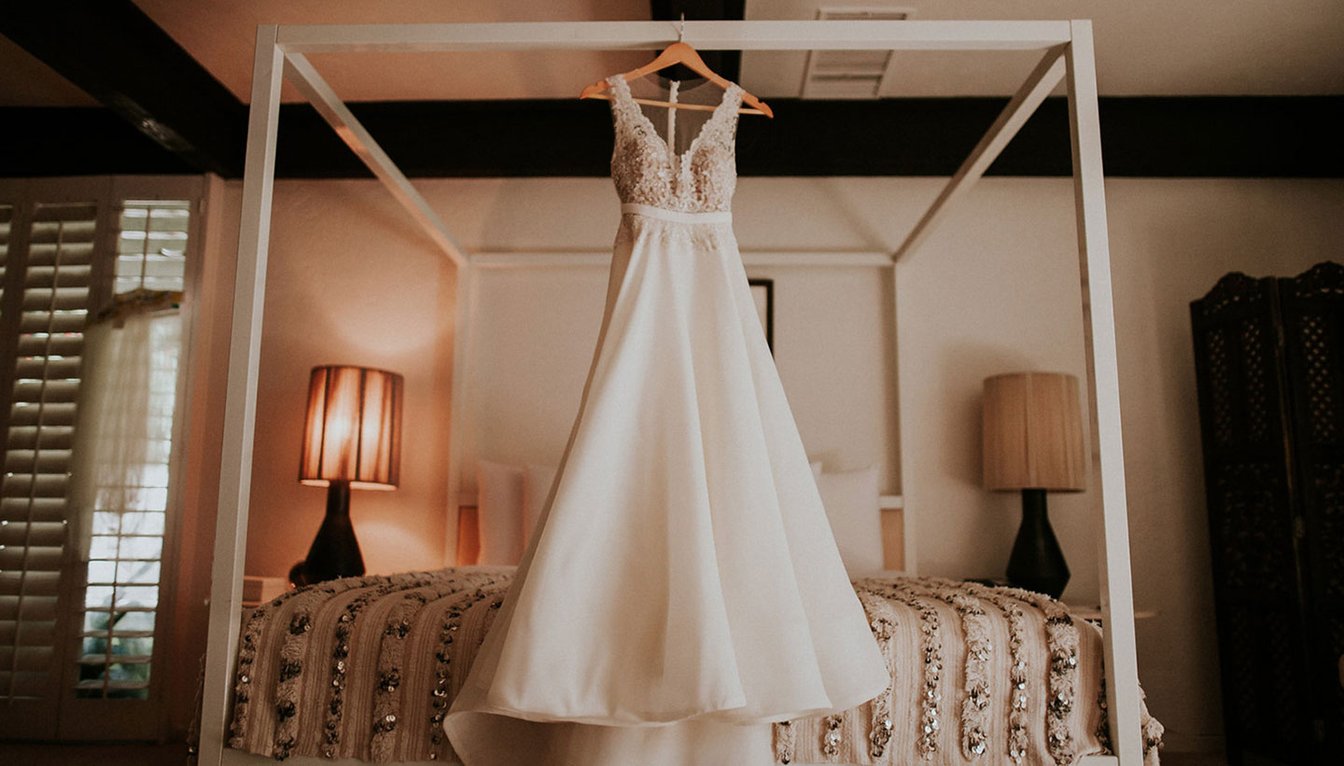 A white wedding dress hangs from a wooden hanger on a white four-poster bed in a room with beige walls and black beams.