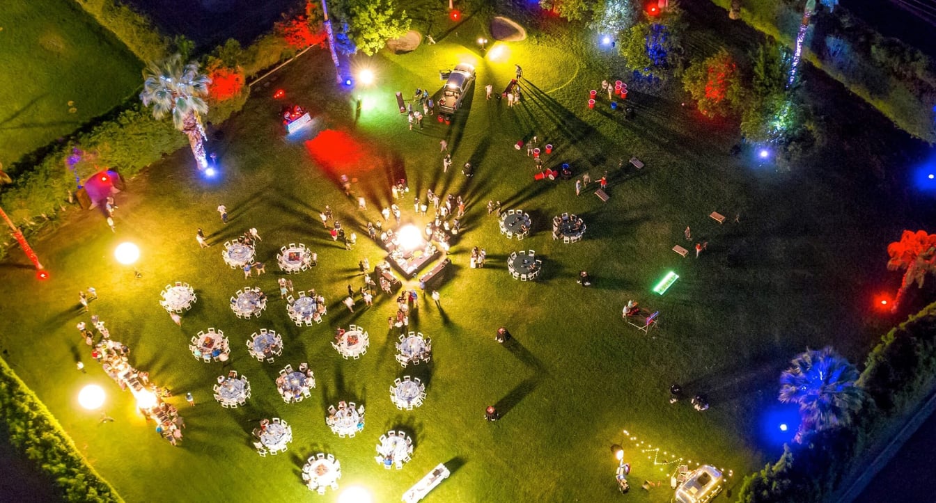 An aerial view of a nighttime outdoor event with tables, chairs, and people on the grass.