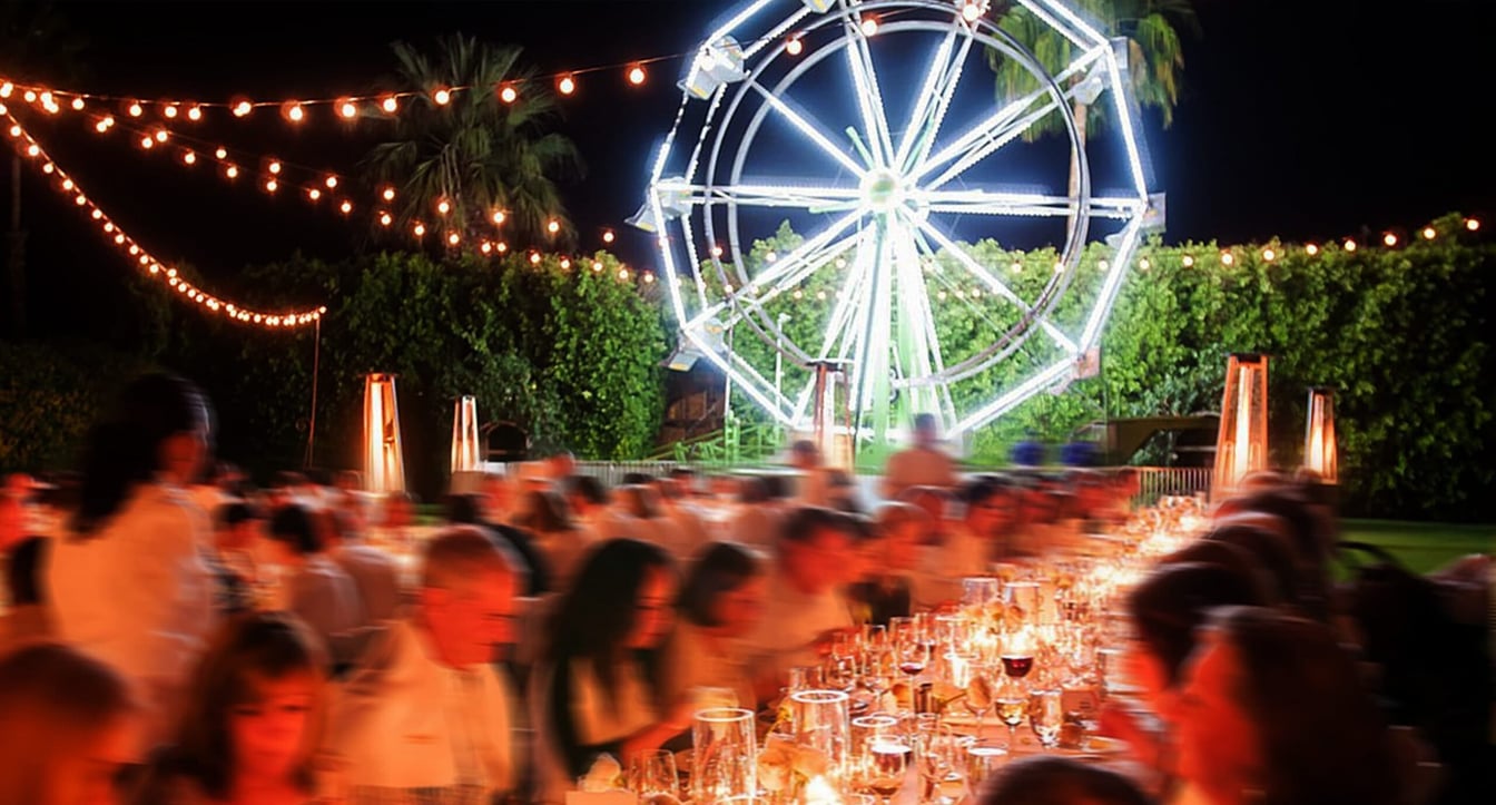 A nighttime outdoor dining event with a large crowd seated at tables, a lit Ferris wheel in the background, and string lights overhead.
