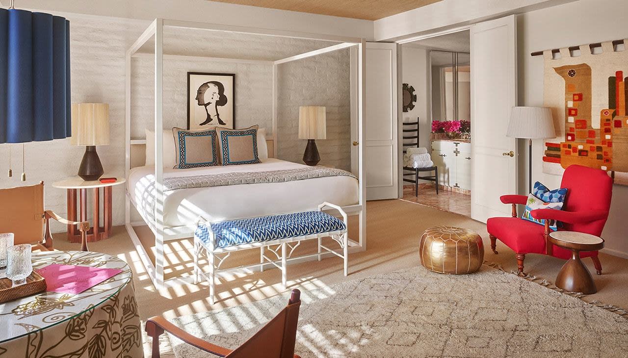 A wide view of a hotel room with a white four-poster bed, blue accents, and a red chair.
