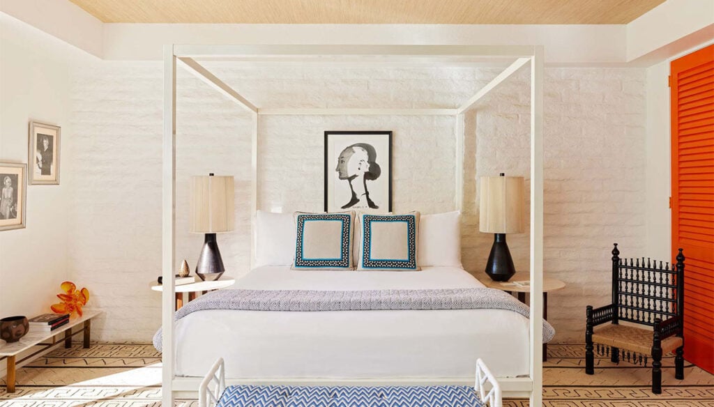 A wide view of a guest room with a four-poster bed, lamps, artwork, chair, and bench.