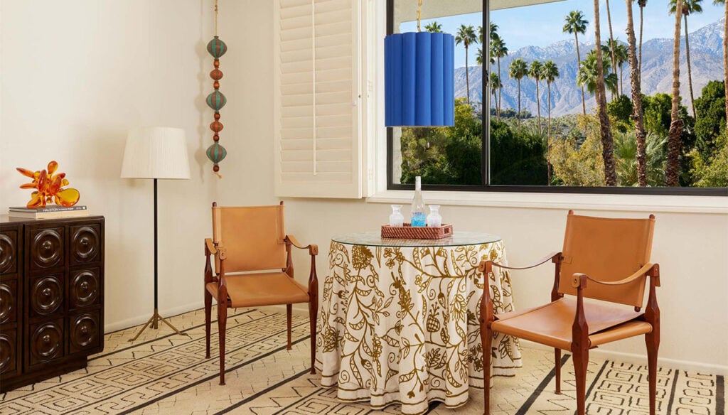 A stylish guest room with a glass table, two leather chairs, and a view of palm trees and mountains outside the window.