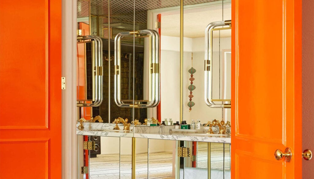 A bathroom sink area with a large mirror, marble countertop, and gold faucets, framed by orange doors.
