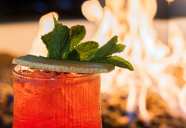 a red cocktail with ice, garnished with cucumber and mint, in front of a fire