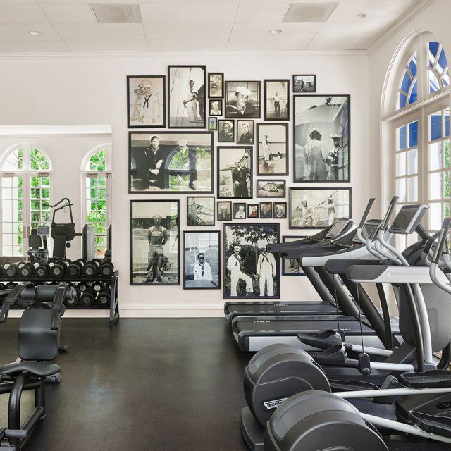 A gym with treadmills, dumbbells, and exercise equipment in front of a wall of framed photos and arched windows.
