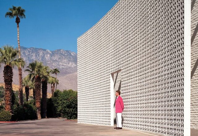A large white building with a unique facade, a person in a pink jacket standing in front of it, palm trees and mountains in the background.