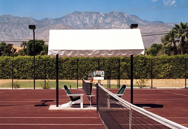 A tennis court with a white awning, surrounded by a black fence and mountains in the background.