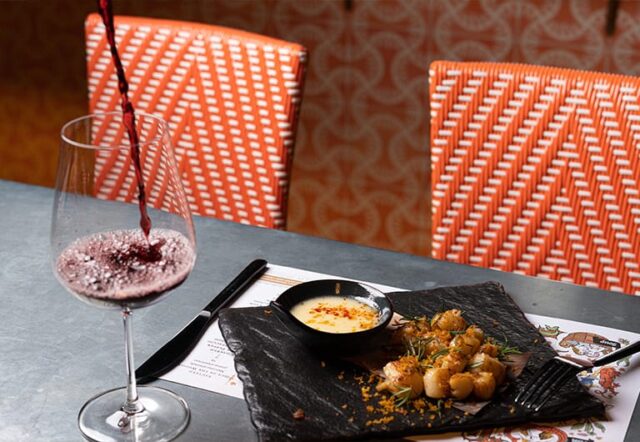 A dining table set with a glass of wine, a plate of food, and a bowl of sauce.