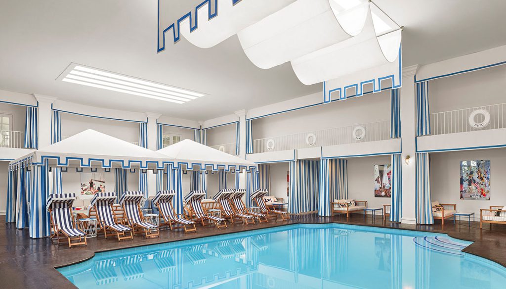 A large indoor pool with lounge chairs and canopies, surrounded by a balcony with blue and white stripes.