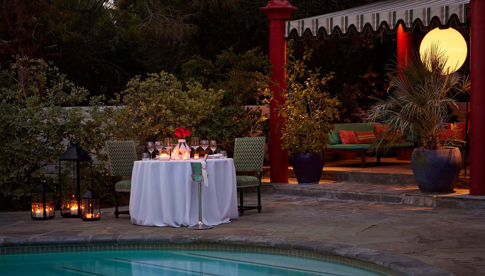 A romantic dinner setup by the pool with a table, chairs, candlelight, flowers, and wine glasses.