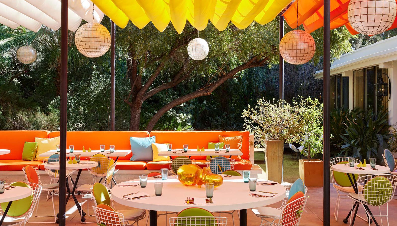 A brightly lit outdoor dining area with colorful tables and chairs, surrounded by trees and greenery.