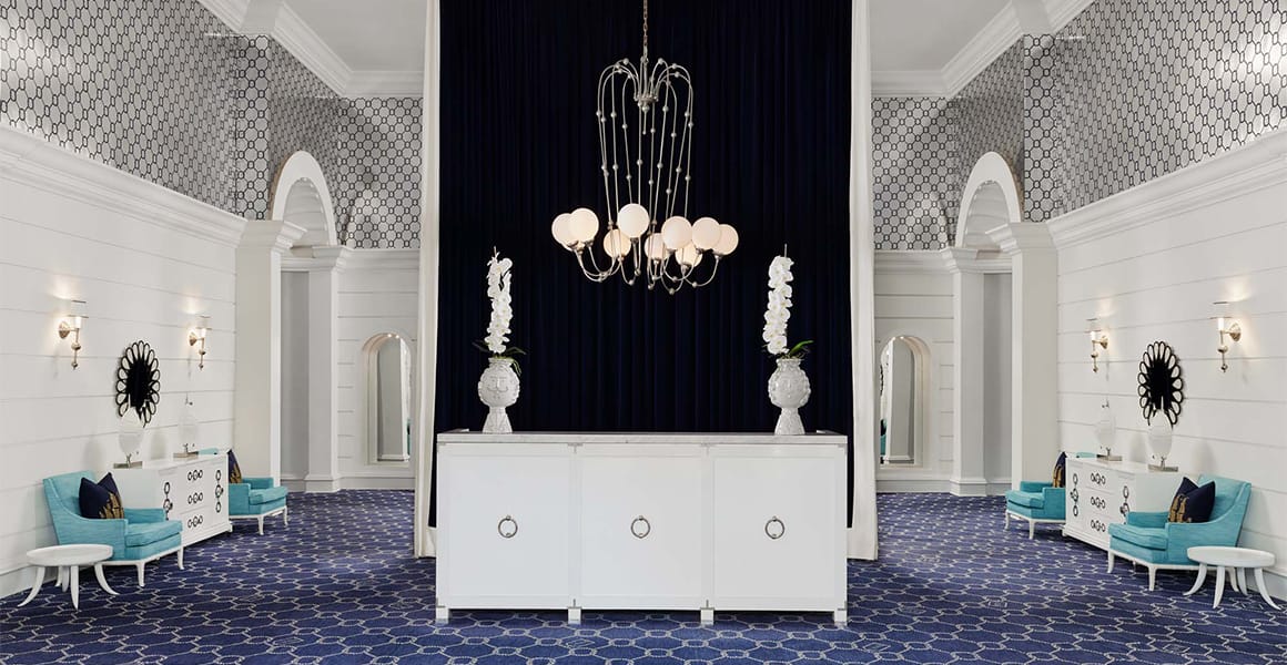 A wide view of a luxurious lobby with a chandelier, blue carpet, and white furniture.