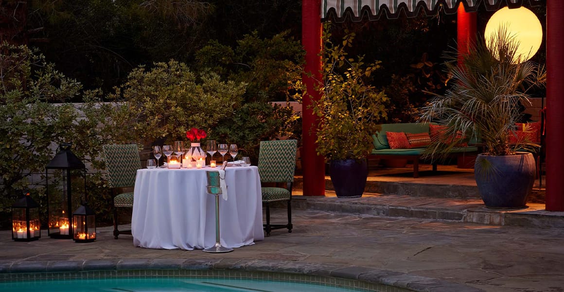 An outdoor dining area by a pool at night with a table set for two, surrounded by lanterns and plants.
