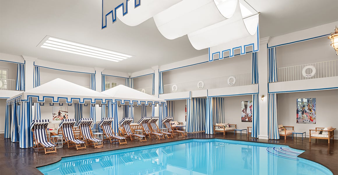 A large indoor pool with lounge chairs and cabanas, surrounded by balcony and artwork.