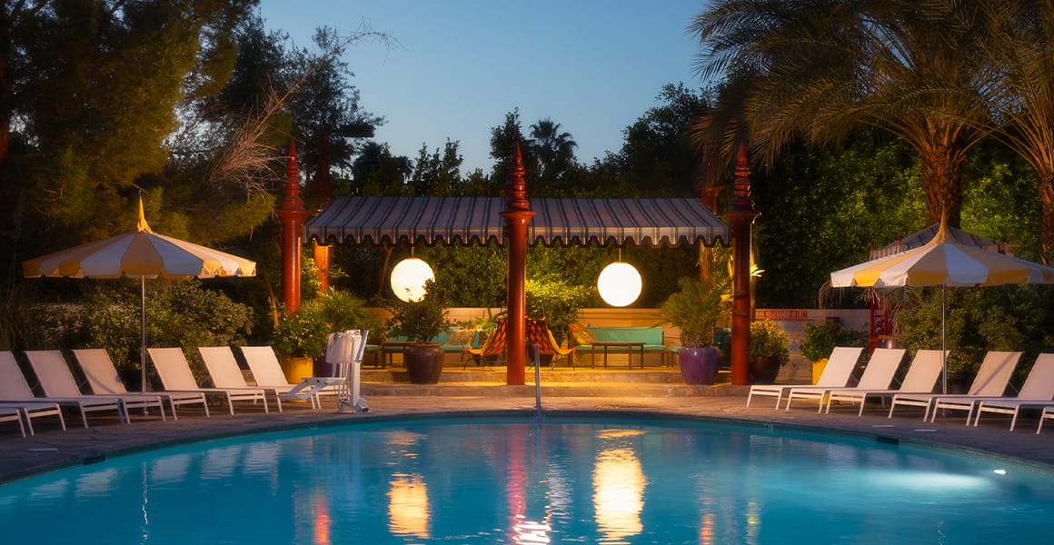 A serene poolside scene at dusk with lounge chairs, umbrellas, and a pool surrounded by trees and a gazebo.