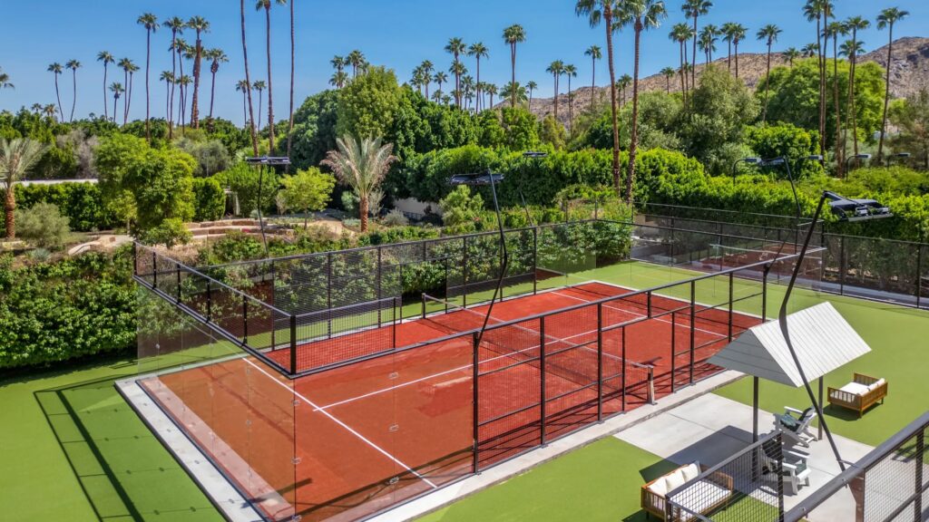 A padel court with a red surface and black fence, surrounded by greenery and palm trees, with a mountain view in the background.