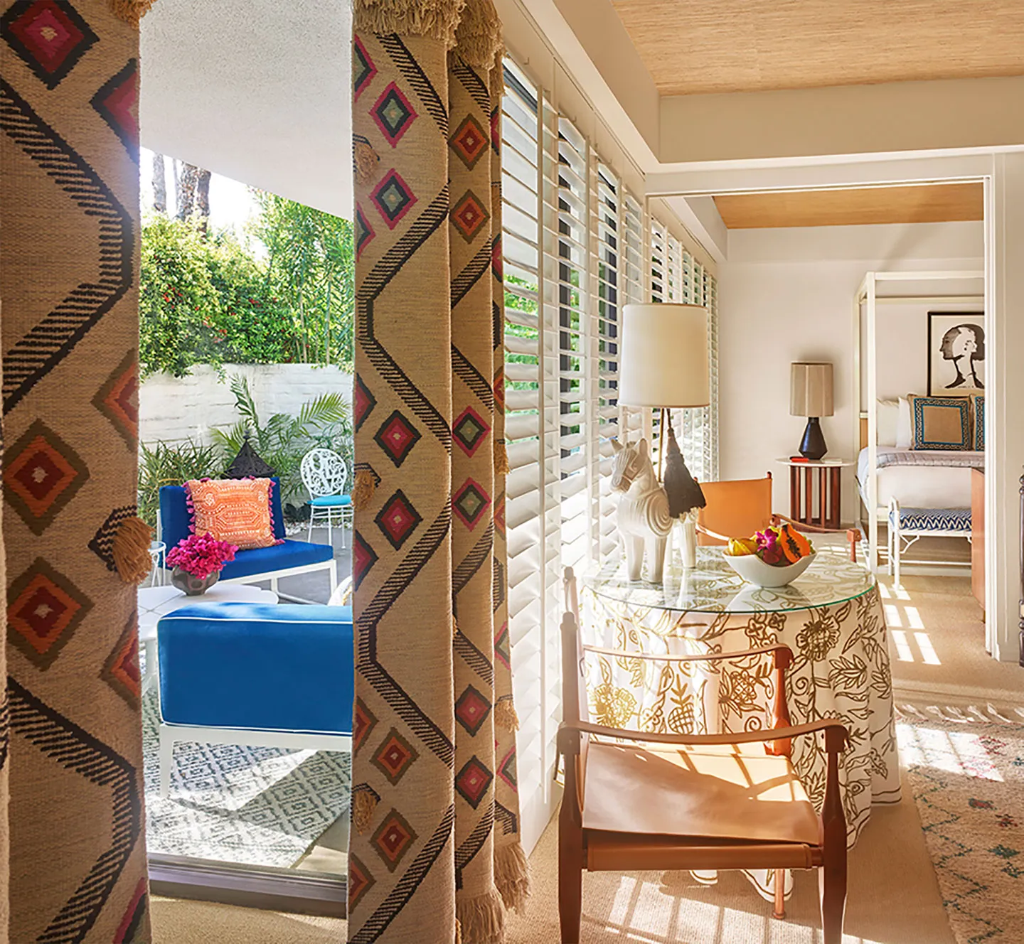 a bright and stylish interior space with floor-to-ceiling windows and white shutters. Inside, there’s a round glass table with a floral patterned cloth, a leather chair, and a ceramic horse lamp. The outdoor patio is visible through the windows, featuring blue cushioned seating, a vibrant orange pillow, and lush greenery