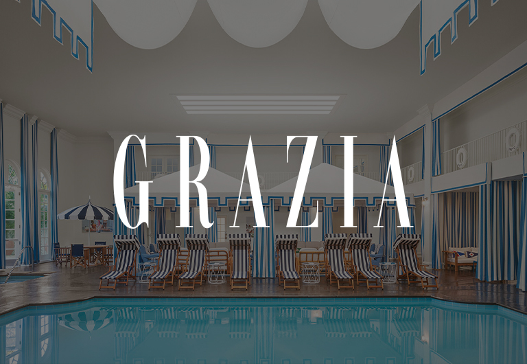 Indoor pool area with striped lounge chairs, umbrellas, and blue and white decor. Large windows line the wall. The text GRAZIA is prominently displayed over the image.