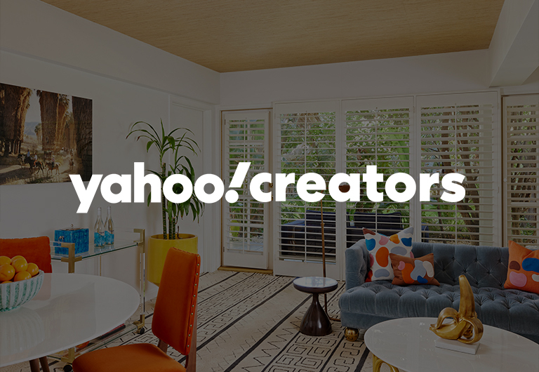 Bright, modern living room with a sofa, vibrant pillows, a small round table, orange chair, fruit bowl, and large windows with blinds. yahoo! creators is written in bold white text across the image.