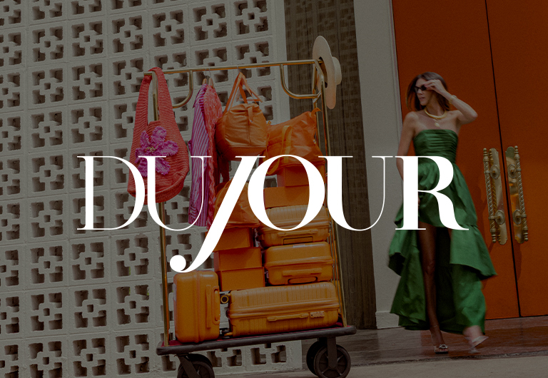A woman in a green dress walks past orange luggage and bags stacked on a hotel cart; the word DUJOUR is overlaid in large white letters across the image.