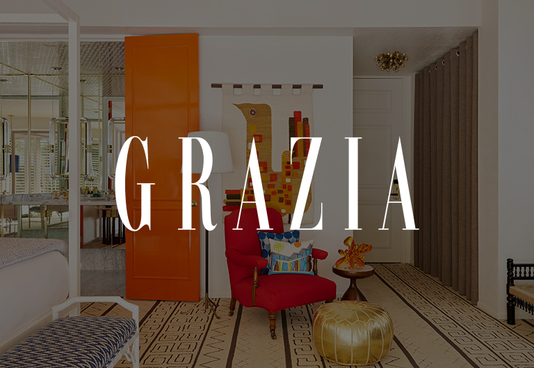 A stylish living room with a red chair, gold ottoman, orange door, geometric wall art, and patterned rug. The word GRAZIA is overlaid in large white letters.
