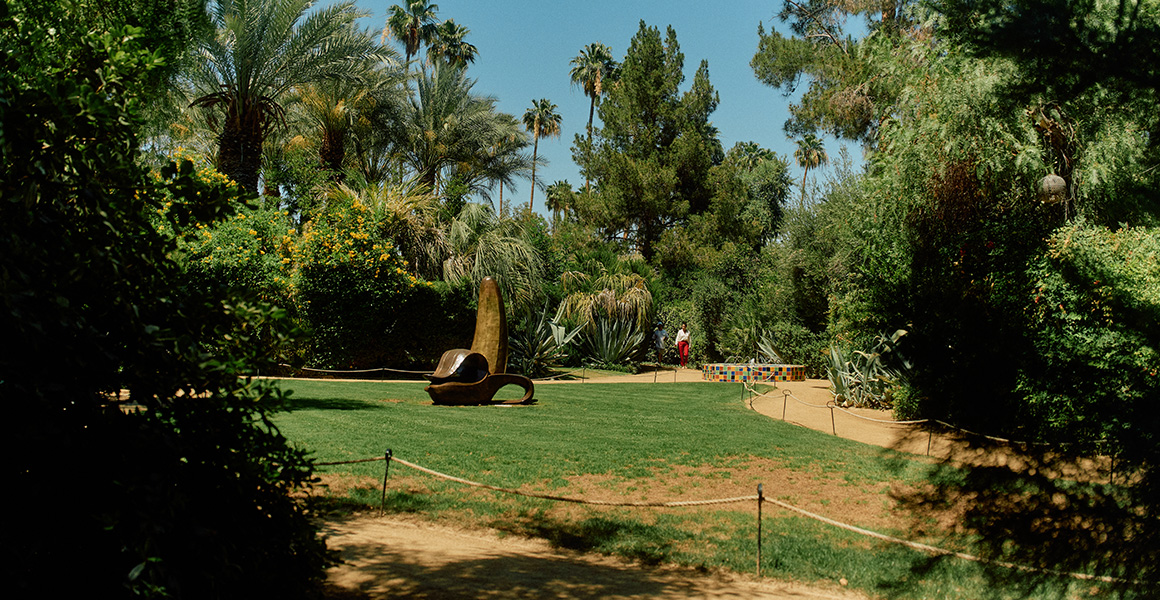 A modern abstract sculpture shaped like a stylized banana sits on a lawn in a garden surrounded by trees and palms.