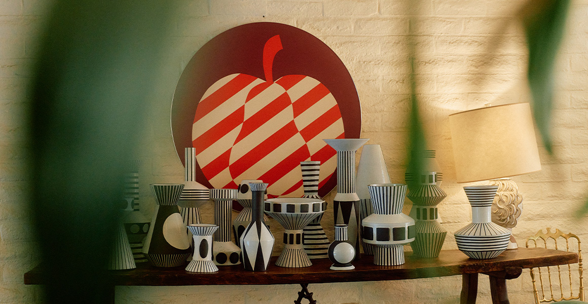 A collection of black-and-white patterned ceramic vases arranged on a wooden shelf in front of a circular red-and-white striped apple art piece.