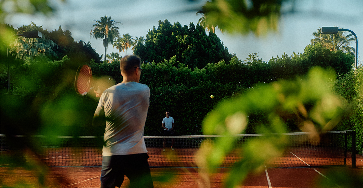 Two men play tennis on an outdoor clay court framed by lush foliage.