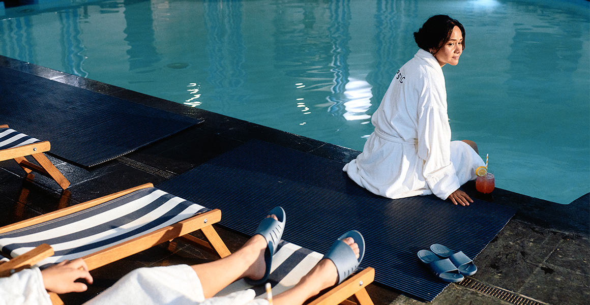 A woman in a white bathrobe sits on the edge of an indoor pool next to a cocktail, while another person wearing blue slide sandals rests on a lounge chair in the foreground.