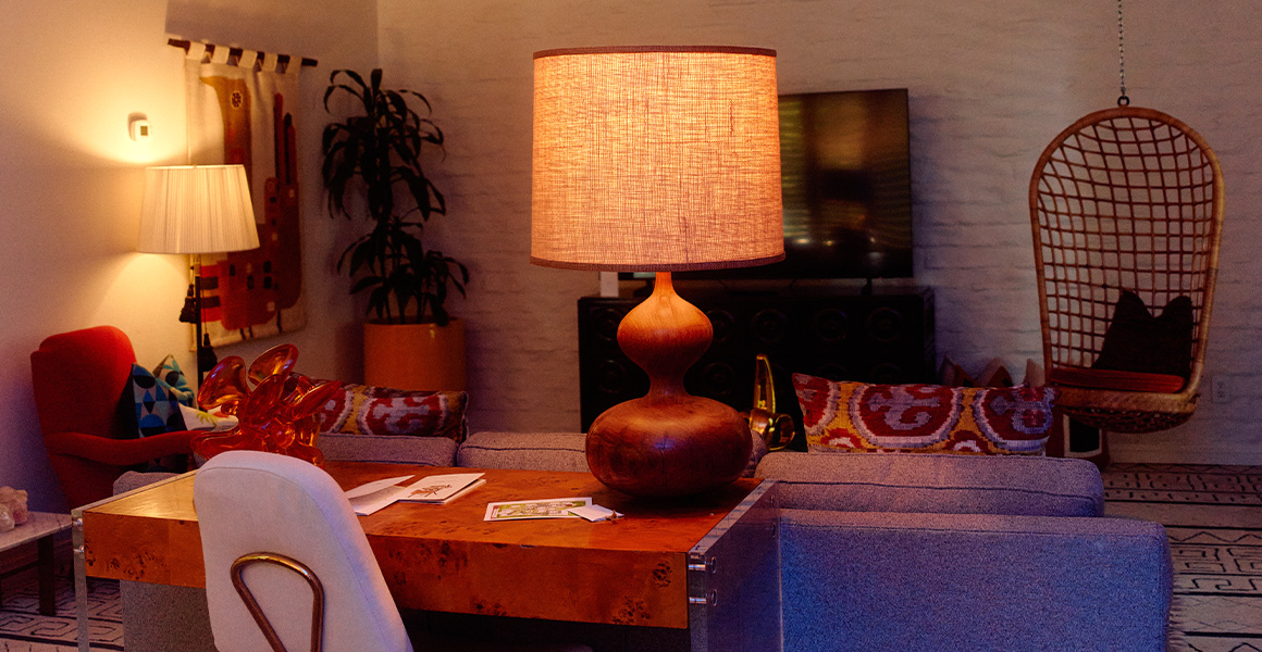 A living room featuring a wooden desk lamp in the foreground, a gray sofa, a hanging rattan chair, and a television against a brick wall.