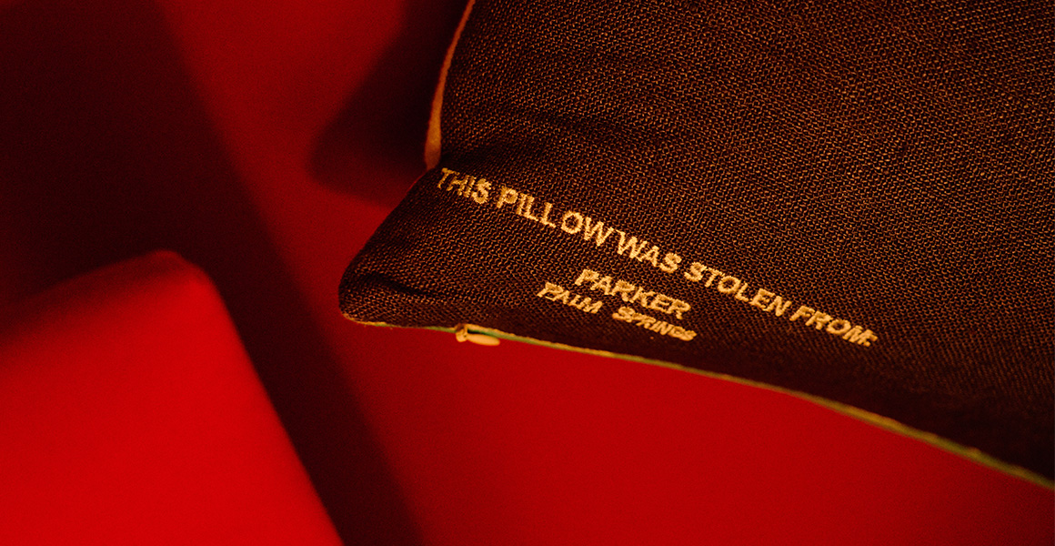A dark pillow with text embroidered in gold that reads, THIS PILLOW WAS STOLEN FROM PARKER PALM SPRINGS.