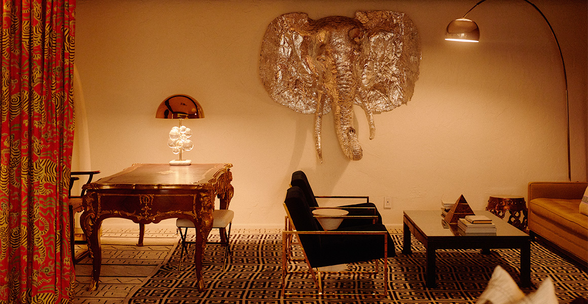 A room featuring an ornate wooden desk with a dome lamp, two black armchairs, a low coffee table, and a large silver elephant head wall sculpture.